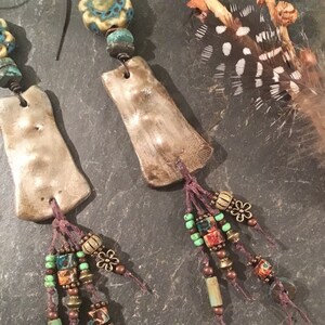 May include: A pair of handmade earrings with a rustic, bohemian style. Each earring features a textured, light brown clay pendant with a turquoise and white floral bead, and a tassel of brown, turquoise, and red beads.