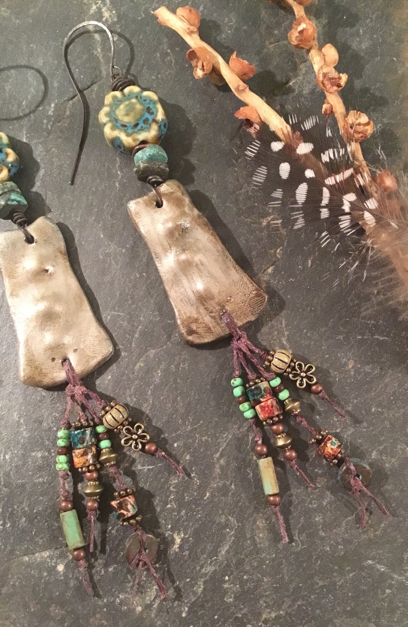 May include: A pair of handmade earrings with a rustic, bohemian style. Each earring features a textured, light brown clay pendant with a turquoise and white floral bead, and a tassel of brown, turquoise, and red beads.