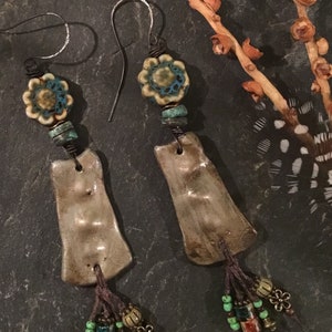 May include: A pair of handmade ceramic earrings with a brown and white glaze. The earrings feature a turquoise and white floral bead, brown beads, and a brown tassel.