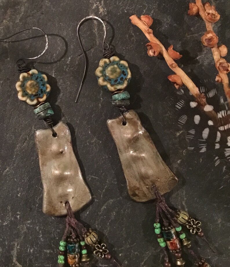 May include: A pair of handmade ceramic earrings with a brown and white glaze. The earrings feature a turquoise and white floral bead, brown beads, and a brown tassel.