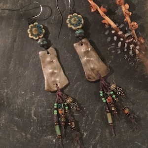 May include: A pair of handmade dangle earrings with brown ceramic teardrop shapes, turquoise and green beads, and brown tassels. The earrings have silver hooks and a turquoise and white floral bead at the top.