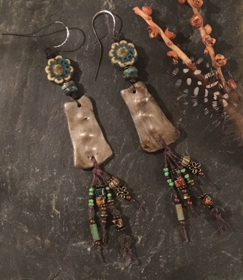 May include: A pair of handmade dangle earrings with brown ceramic teardrop shapes, turquoise and green beads, and brown tassels. The earrings have silver hooks and a turquoise and white floral bead at the top.