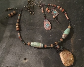 May include: A multi-strand necklace with a large, light brown pendant and a smaller, turquoise pendant. The necklace is made of various beads, including brown, turquoise, and gold. The necklace is on a dark gray background.