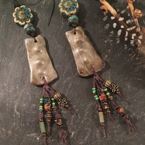 May include: A pair of handmade earrings with a rustic, bohemian style. Each earring features a silver-toned clay pendant with a textured surface, a turquoise and white floral bead, and a tassel of brown cord and colorful beads.