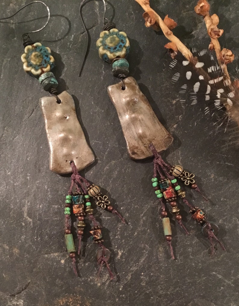 May include: A pair of handmade earrings with a rustic, bohemian style. Each earring features a silver-toned clay pendant with a textured surface, a turquoise and white floral bead, and a tassel of brown cord and colorful beads.