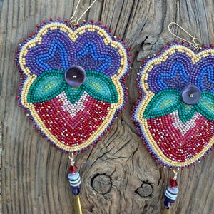 May include: A pair of beaded earrings shaped like strawberries with purple, blue, green, red, and yellow seed beads. Each earring features a dark purple button center, a gold-tone hook, and a dangling tassel with red and gray beads.