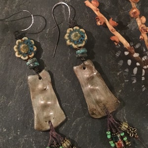 May include: A pair of handmade dangle earrings with brown leather tassels, ceramic beads, and silver hooks. The ceramic beads are a light brown with a blue and white floral design.
