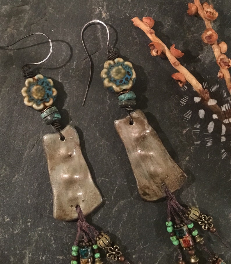 May include: A pair of handmade dangle earrings with brown leather tassels, ceramic beads, and silver hooks. The ceramic beads are a light brown with a blue and white floral design.