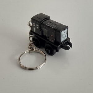 May include: A black toy train engine keychain with a silver key ring. The train has a smiling face and is detailed with windows and other features. The keychain is a miniature replica of a train engine.