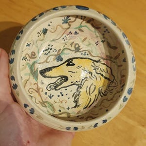 以下が含まれることがあります： 黄色と黒で犬の頭が手描きされた白い陶器のボウル。ボウルは青と白の花柄の縁取りがされています。