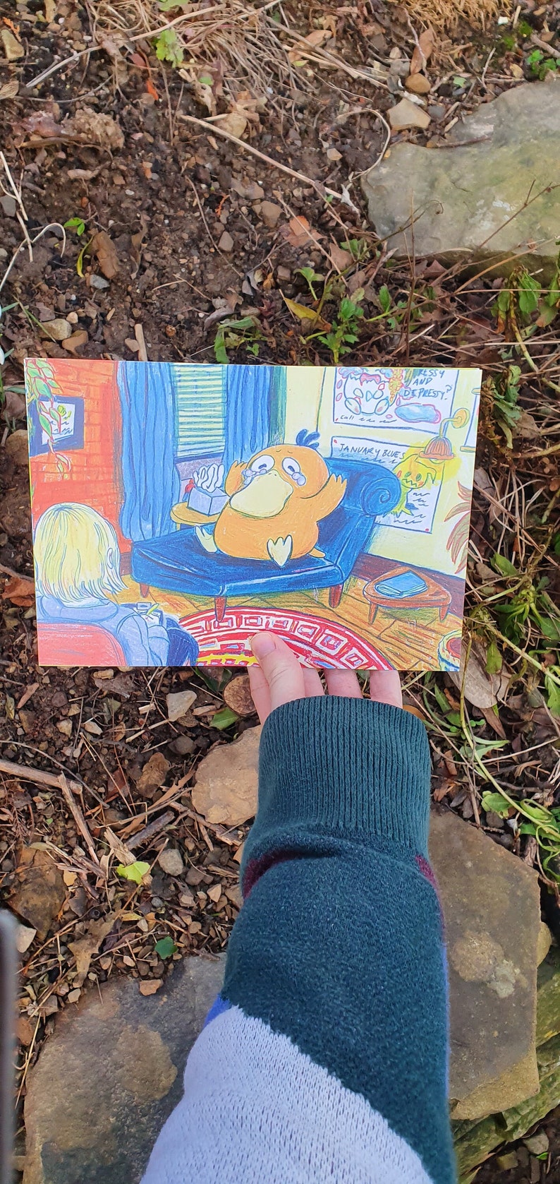 May include: A hand holds a cartoon illustration of a yellow duck-like creature sitting on a blue couch. The duck is crying and has a box of tissues next to it. The text on the wall reads "January Blues" and "Are you okay, my dear?"