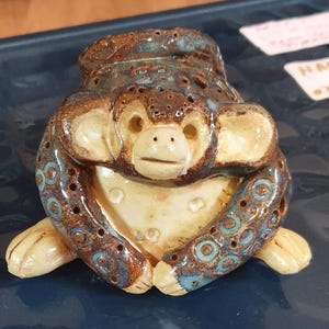 May include: A ceramic monkey figurine with a brown and cream color scheme, accented with blue details. The monkey has a smiling face, large ears, and is posed with its hands together. The figurine has a textured surface and is likely handmade.