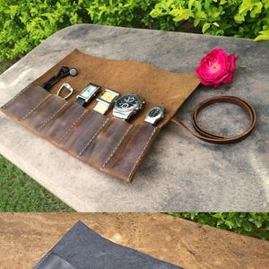 May include: A brown leather watch roll with five compartments, holding several wristwatches. The roll is open, revealing the watches, and has a leather strap for closure. A pink rose is in the background.