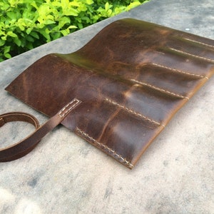 May include: A brown leather roll-up case with white stitching. The case has multiple compartments and a leather strap for securing it. The leather has a distressed look.