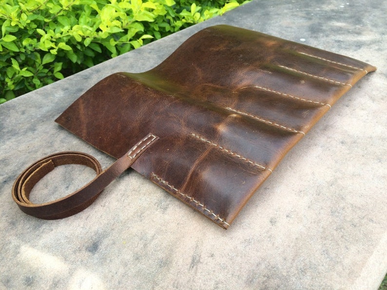 May include: A brown leather roll-up case with white stitching. The case has multiple compartments and a leather strap for securing it. The leather has a distressed look.