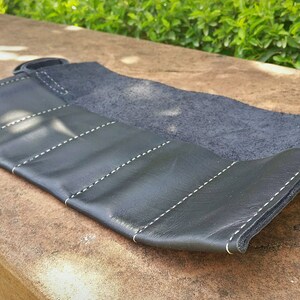 May include: Black leather roll-up case with white stitching. The case is partially unrolled, revealing a dark interior. The exterior has multiple sections, each stitched with a white thread. The case is designed to hold items.