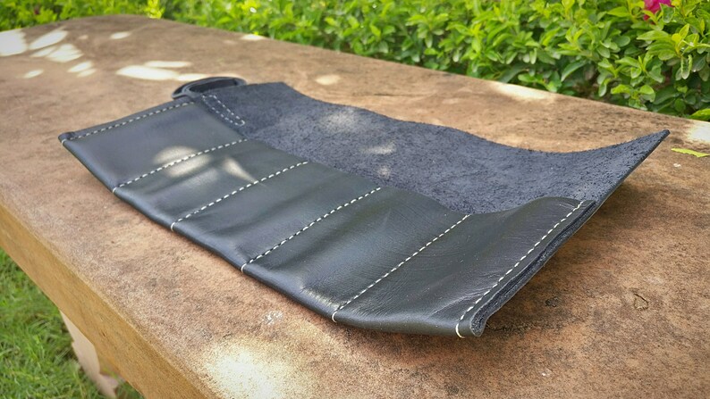 May include: Black leather roll-up case with white stitching. The case is partially unrolled, revealing a dark interior. The exterior has multiple sections, each stitched with a white thread. The case is designed to hold items.