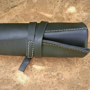 May include: A rolled-up, dark green leather pencil case with a strap closure. The case is secured with a matching leather strap that wraps around the body. White stitching accents the edges, and the case is set against a textured, light brown surface.