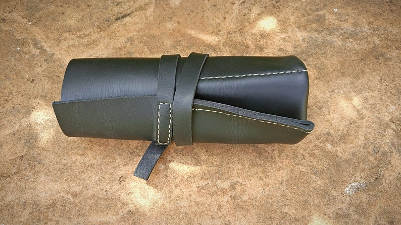 May include: A rolled-up, dark green leather pencil case with a strap closure. The case is secured with a matching leather strap that wraps around the body. White stitching accents the edges, and the case is set against a textured, light brown surface.