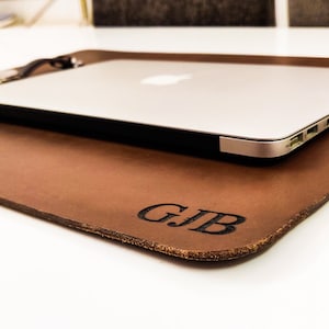 Puede incluir: Un tapete de escritorio de cuero marrón con un ordenador portátil encima. El tapete tiene un aspecto envejecido y las iniciales "GJB" están grabadas en la esquina inferior derecha. Un ordenador portátil plateado está colocado sobre el tapete.