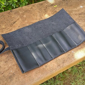 May include: A black leather roll-up case with white stitching. The case has multiple compartments and a strap for securing it. The case is open, revealing the interior. The background is a natural setting with greenery.