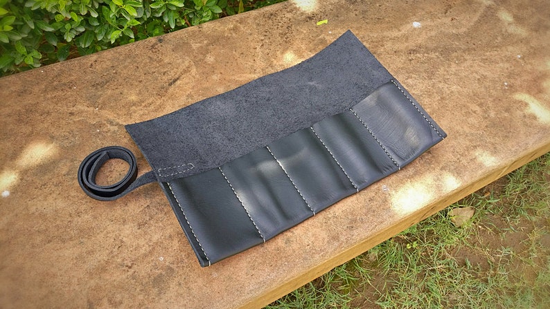 May include: A black leather roll-up case with white stitching. The case has multiple compartments and a strap for securing it. The case is open, revealing the interior. The background is a natural setting with greenery.