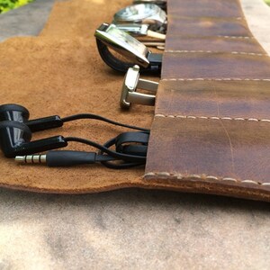 May include: A brown leather watch roll with several watches, a pair of silver cufflinks, and black earbuds. The roll is open, revealing the items inside. The leather has a distressed look with white stitching.