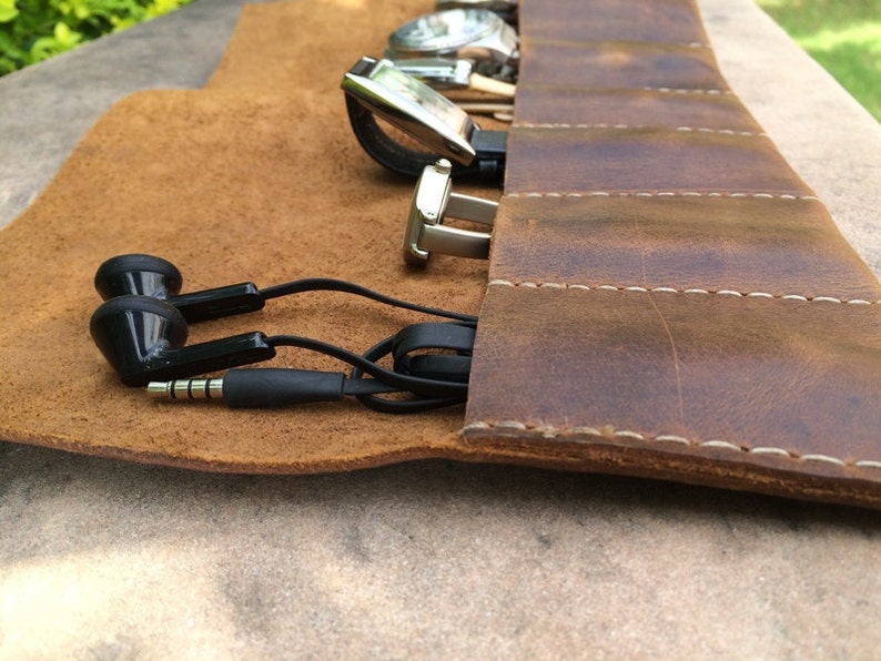 May include: A brown leather watch roll with several watches, a pair of silver cufflinks, and black earbuds. The roll is open, revealing the items inside. The leather has a distressed look with white stitching.