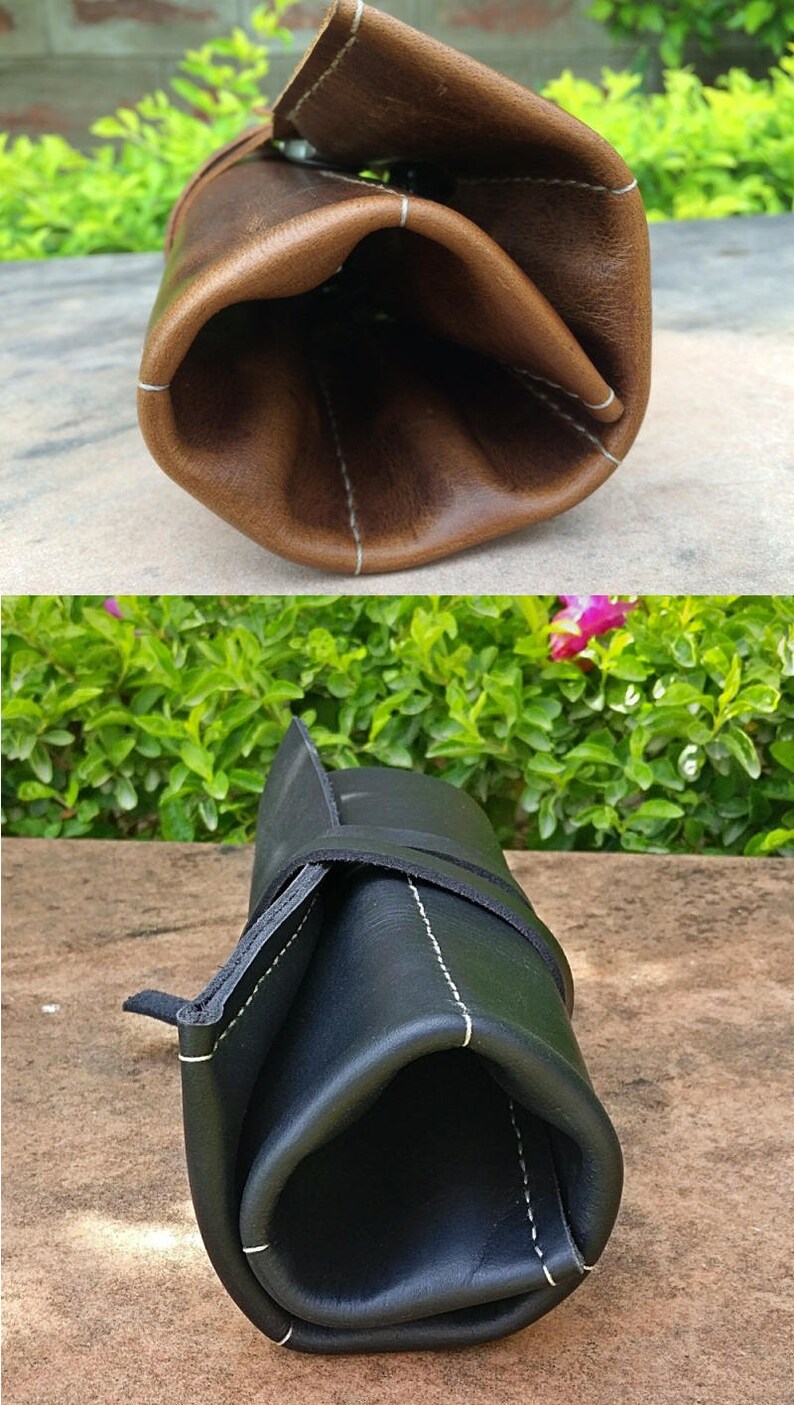 May include: Two leather roll-up cases, one brown and one black, are displayed. The brown case is open, revealing its interior, while the black case is closed and secured with a strap. Both cases have white stitching.