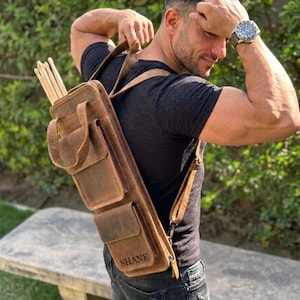 Puede incluir: Un carcaj de cuero marrón con múltiples bolsillos y correas para los hombros. El carcaj está personalizado con el nombre "SHANE" y contiene varias flechas de madera. El diseño incluye un asa y un cierre de hebilla. El carcaj se lleva sobre el hombro.