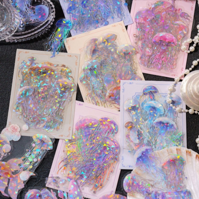May include: A set of iridescent holographic stickers featuring jellyfish in various poses. The stickers are on a white background with a delicate floral border.