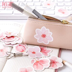 May include: A set of pink and white watercolor flower stickers. The stickers feature a delicate floral design with pink and white petals and a red center. The stickers are arranged in a pile on a white notebook with a pencil and a silver binder clip.