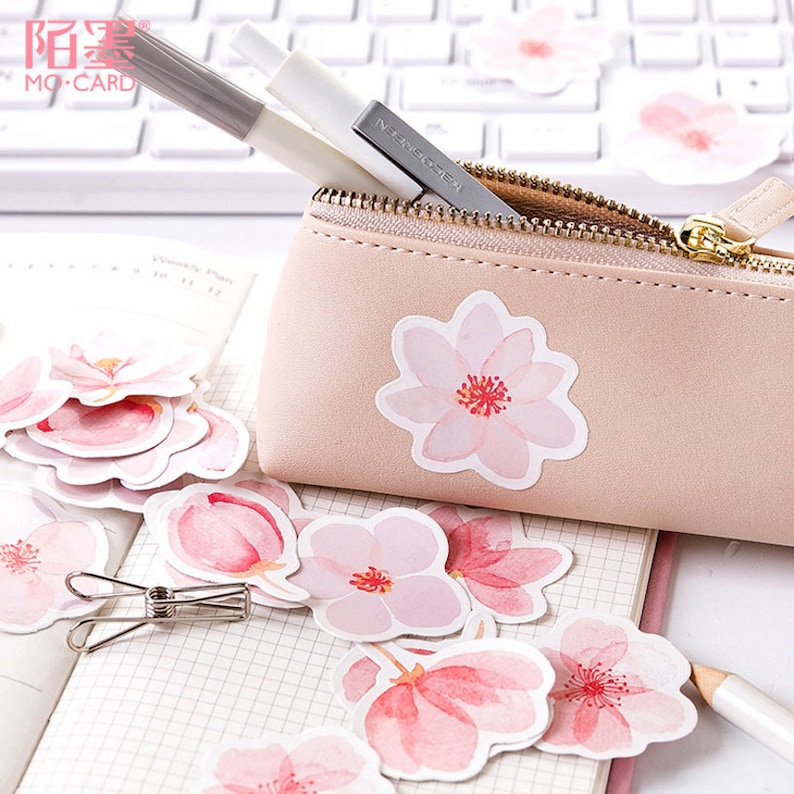 May include: A set of pink and white watercolor flower stickers. The stickers feature a delicate floral design with pink and white petals and a red center. The stickers are arranged in a pile on a white notebook with a pencil and a silver binder clip.