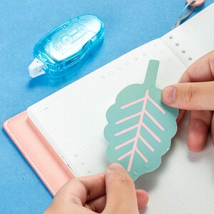 May include: A person is using a blue tape dispenser to attach a green leaf-shaped paper sticker to a pink and white notebook. The sticker has a pink vein pattern.