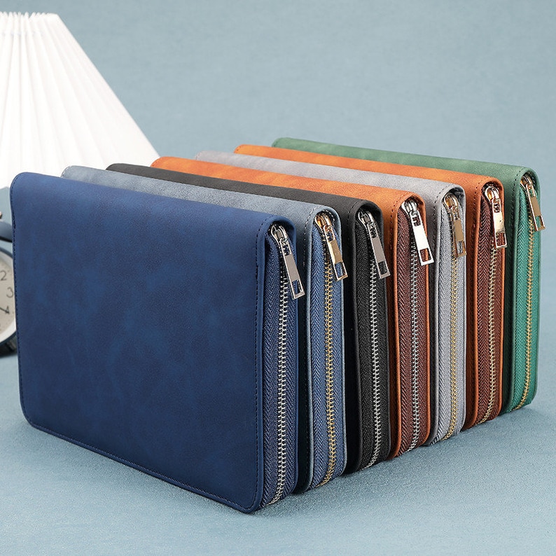 K&ouml;nnte beinhalten: Eine Reihe von Dokumentenhaltern mit Rei&szlig;verschluss in verschiedenen Farben, darunter Blau, Schwarz und Braun. Jeder Halter hat einen goldfarbenen Rei&szlig;verschluss und eine rechteckige Form. Die Halter sind diagonal angeordnet und zeigen ihre verschiedenen Farbt&ouml;ne.