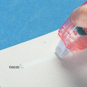 May include: A clear, pink tape dispenser with a white roller is dispensing a 6mm wide strip of white tape onto a gridded sheet of paper.