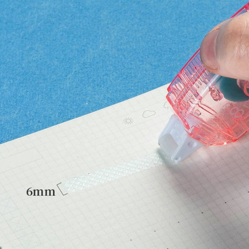 May include: A clear, pink tape dispenser with a white roller is dispensing a 6mm wide strip of white tape onto a gridded sheet of paper.