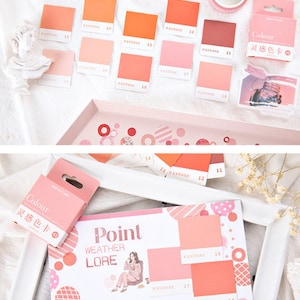 May include: A set of color swatches with the word "PANTONE" and numbers printed on each swatch. The swatches are arranged in a grid pattern on a white surface. The swatches are in shades of pink, orange, and red. The text "Colour 灵感色卡" is printed on a small box.