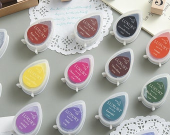 Puede incluir: Una colección de almohadillas de tinta en forma de lágrima en varios colores, incluyendo naranja, morado y verde. Cada almohadilla está etiquetada con el nombre del color y la palabra "LUXURY". Las almohadillas de tinta están en pequeños recipientes de plástico transparente.