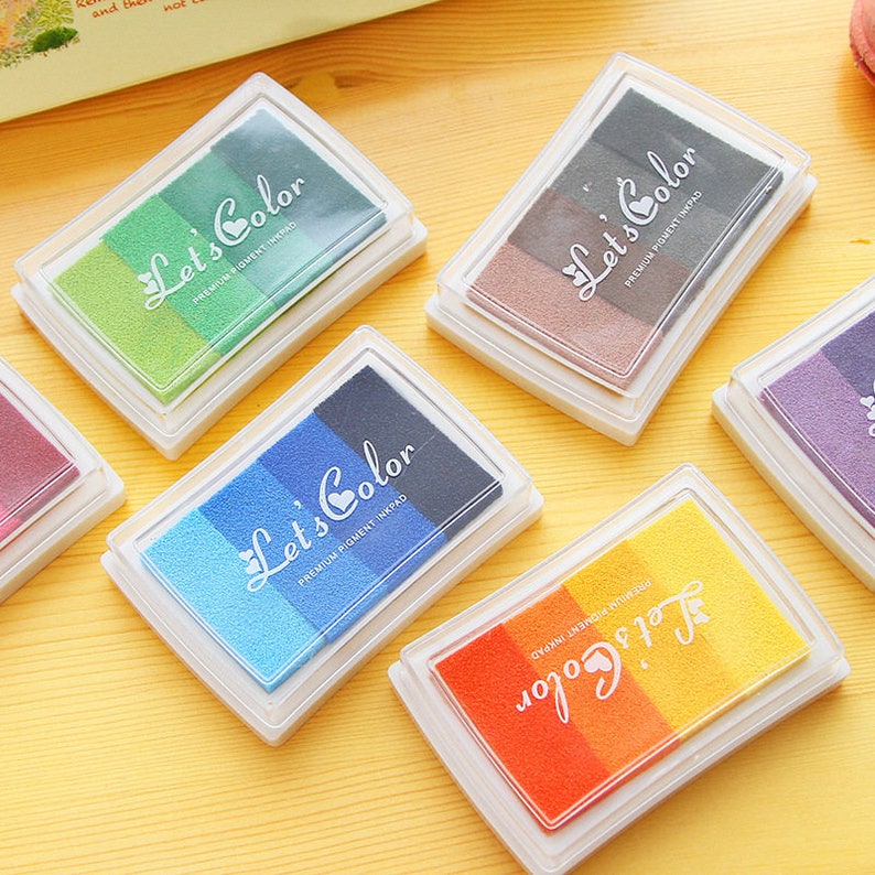 May include: Six ink pads with different color combinations. Each pad has a clear plastic case and a label that says "Let's Color" with a heart symbol. The ink pads are arranged on a yellow wooden surface.