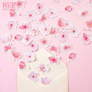 May include: Pink watercolor stickers featuring various pink flowers, including cherry blossoms, with white outlines. The stickers are scattered on a pink background and some are inside a white envelope.