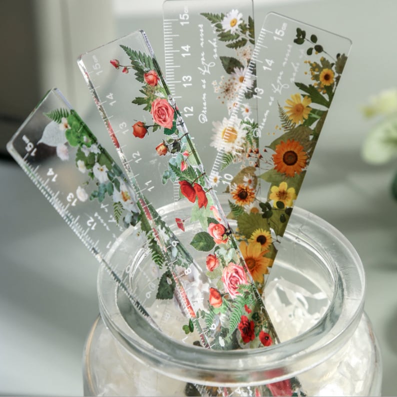 May include: Four clear plastic rulers with floral designs and black numbers. The rulers are 15 inches long and have measurements in inches. The floral designs include roses, daisies, and other flowers.