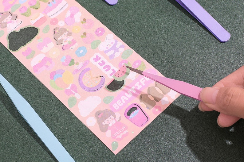 Op de afbeelding: Een vel met roze en witte stickers met verschillende schattige ontwerpen, waaronder een watermeloen, een meisje met een bloemenkrans en een donut. De stickers worden opgepakt met een roze pincet.