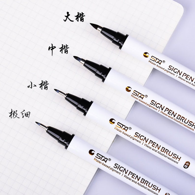 以下が含まれることがあります： 白いバレルと金色の文字で書かれた4本の黒い筆ペン。ペンには「STA SIGN PEN BRUSH」とラベルが貼られており、異なるサイズのチップが付いています。白い紙に黒いインクで「大楷」「中楷」「小楷」「極細」と書いてあります。