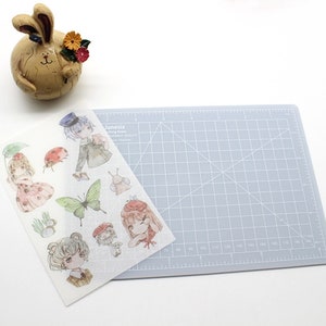 K&ouml;nnte beinhalten: Ein Blatt transparentes Stickerpapier mit einer Vielzahl von niedlichen Comicfiguren und Illustrationen. Die Figuren umfassen ein M&auml;dchen mit einem Marienk&auml;fer, ein M&auml;dchen mit einem Regenschirm, ein Junge mit einem Hut, ein Schmetterling, eine Schnecke, ein Pilz und ein B&auml;r. Das Stickerpapier liegt auf einer grauen Schneidematte.