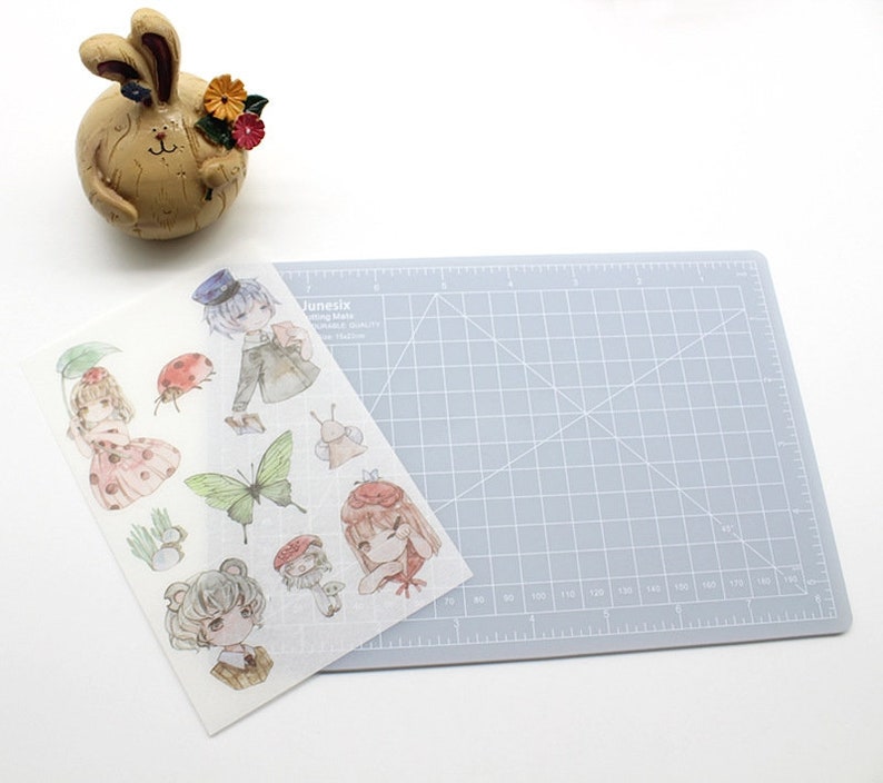 K&ouml;nnte beinhalten: Ein Blatt transparentes Stickerpapier mit einer Vielzahl von niedlichen Comicfiguren und Illustrationen. Die Figuren umfassen ein M&auml;dchen mit einem Marienk&auml;fer, ein M&auml;dchen mit einem Regenschirm, ein Junge mit einem Hut, ein Schmetterling, eine Schnecke, ein Pilz und ein B&auml;r. Das Stickerpapier liegt auf einer grauen Schneidematte.