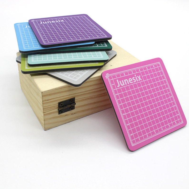 May include: A set of six self-healing cutting mats in various colors, including purple, blue, green, yellow, gray, and pink. The mats are in a wooden box with a hinged lid. Each mat has a grid pattern and the word "Junesix" printed on it.