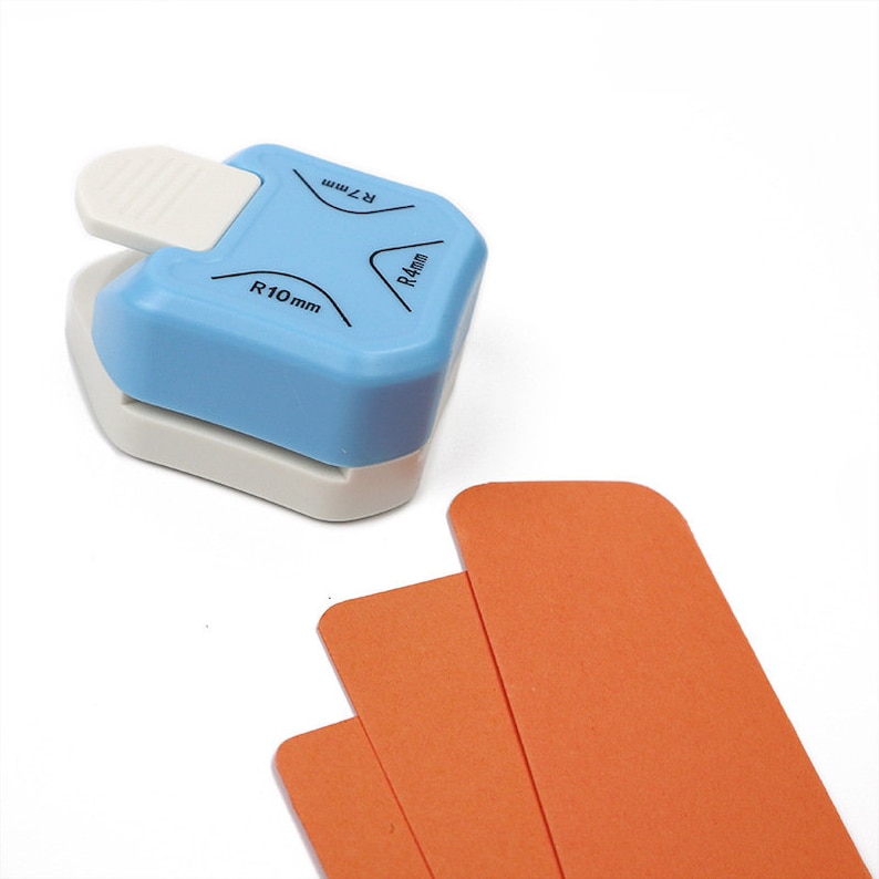 Puede incluir: Un redondeador de esquinas azul y blanco con tres tama&ntilde;os de radio diferentes: R4 mm, R7 mm y R10 mm. El redondeador de esquinas se muestra con tres tiras de papel naranja.
