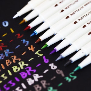 May include: A collection of white metallic pens with various colored tips, including gold, red, and blue, are arranged diagonally on a black surface. The pens are labeled "METILLIC" and are next to colorful handwritten numbers and letters.