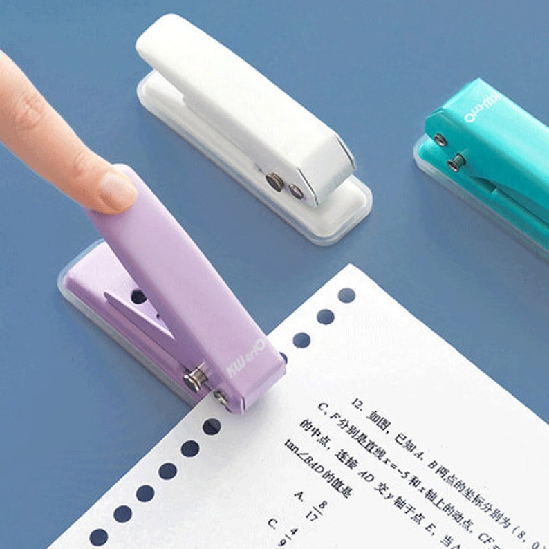 May include: Three hole punchers in different colors: white, purple, and turquoise. The punchers are on a white surface with a sheet of paper with holes punched in it.